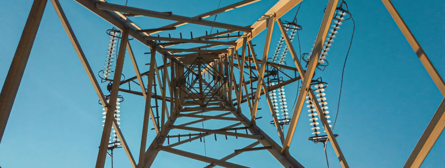 View looking up at the metal framework of an electrical transmission tower against a clear blue sky.