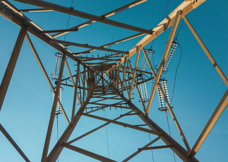View looking up at the metal framework of an electrical transmission tower against a clear blue sky.