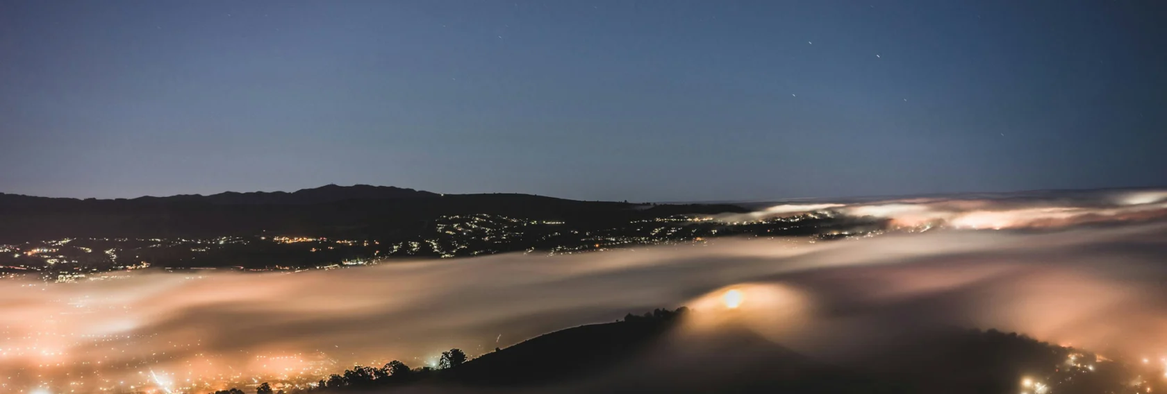 A nighttime cityscape with glowing lights, rolling fog covering the hills, and a clear sky with visible stars above.