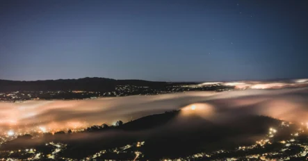 A nighttime cityscape with glowing lights, rolling fog covering the hills, and a clear sky with visible stars above.