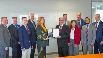 A group of eleven people in business attire stand indoors; two people at the center hold a document together.