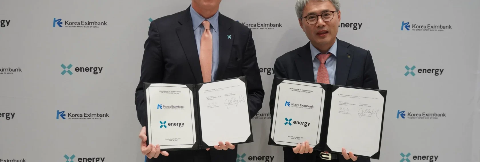 Two men in suits stand in front of a backdrop, holding signed documents from Korea Eximbank and X-energy, indicating a formal agreement or partnership.