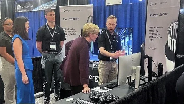 A group of people stands at an exhibition booth, where a man explains a computer display about nuclear reactor technology to a woman. Banners for Fuel: TRISO-X and Reactor: Xe-100 are visible.