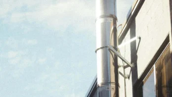 A metal ventilation pipe runs along the exterior wall of a building under a blue sky with scattered clouds.
