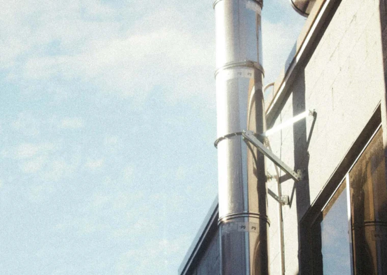 A metal ventilation pipe runs along the exterior wall of a building under a blue sky with scattered clouds.
