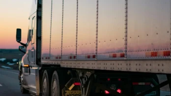 A semi-truck drives on a highway at sunset, with its trailer reflecting the light and the road visible alongside.