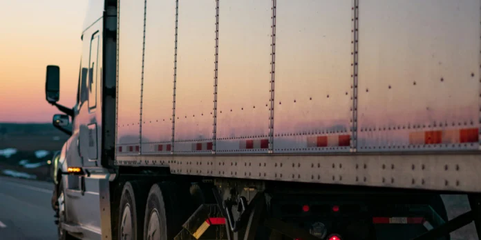 A semi-truck drives on a highway at sunset, with its trailer reflecting the light and the road visible alongside.