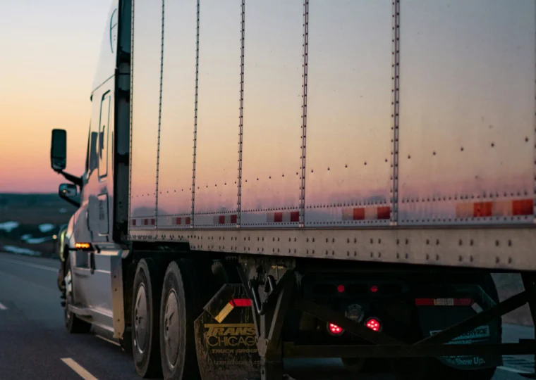 A semi-truck drives on a highway at sunset, with its trailer reflecting the light and the road visible alongside.