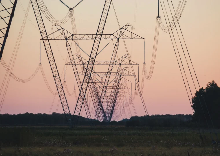 High-voltage transmission towers and power lines stretch across a grassy field at sunset, with trees visible in the background.