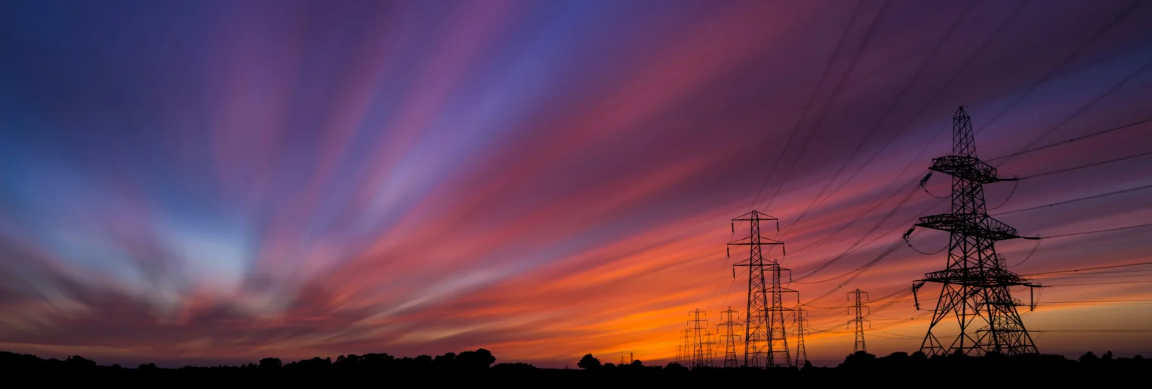 High-voltage power lines and pylons are silhouetted against a colorful sunset sky with streaked clouds.