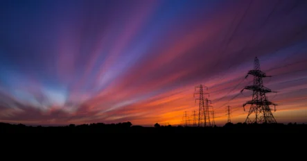 High-voltage power lines and pylons are silhouetted against a colorful sunset sky with streaked clouds.