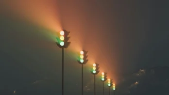 A row of traffic lights with green and amber lights illuminated, casting colorful beams into a dark, misty landscape with mountains in the background.
