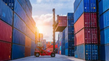 A forklift lifts a shipping container between tall stacks of colorful cargo containers at a shipping yard, with the sun setting in the background.