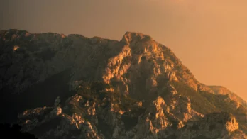 Mountain peaks illuminated by warm sunlight under a partly cloudy sky, with shadows covering the foreground and trees visible at the base.