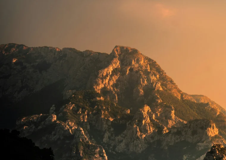 Mountain peaks illuminated by warm sunlight under a partly cloudy sky, with shadows covering the foreground and trees visible at the base.