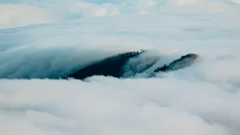 A hilltop covered with trees partially emerges through a dense layer of clouds under a clear sky.