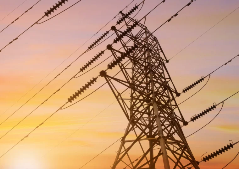 A metal electricity pylon with multiple power lines is silhouetted against a colorful sunset sky.