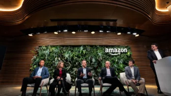 Five people sit on stage in front of a green wall and the Amazon logo; four participate in a panel discussion while one stands at a podium to the right.