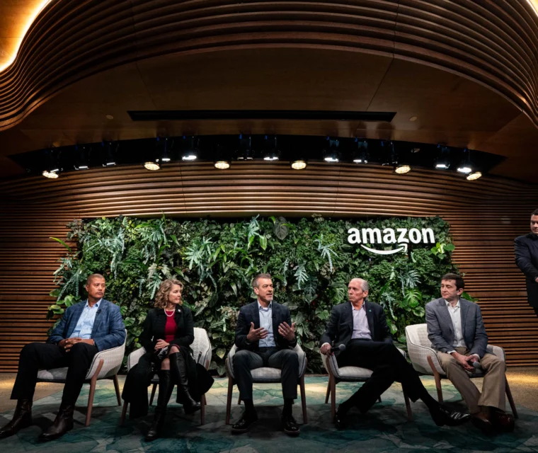 Five people sit on stage in front of a green wall and the Amazon logo; four participate in a panel discussion while one stands at a podium to the right.