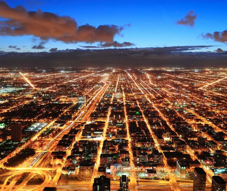 Aerial view of a city at dusk with illuminated streets forming a grid pattern, buildings in the foreground, and a deep blue sky with clouds above.