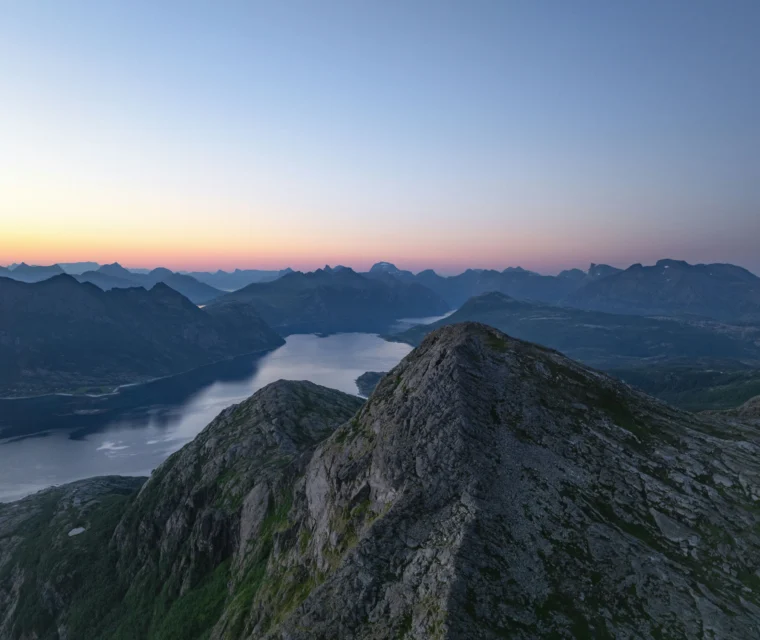 Mountain ridge overlooking a lake and distant mountains at sunset, with a clear sky and soft twilight colors on the horizon.