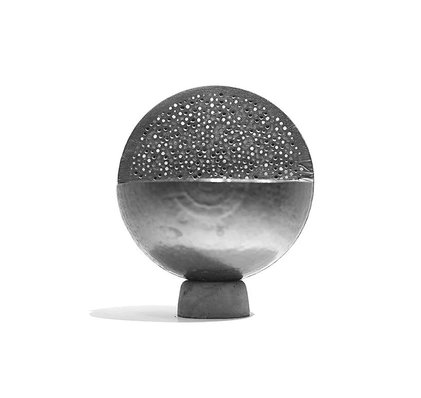 A round metal tea infuser with small holes on the top half, standing upright on a small base against a white background.