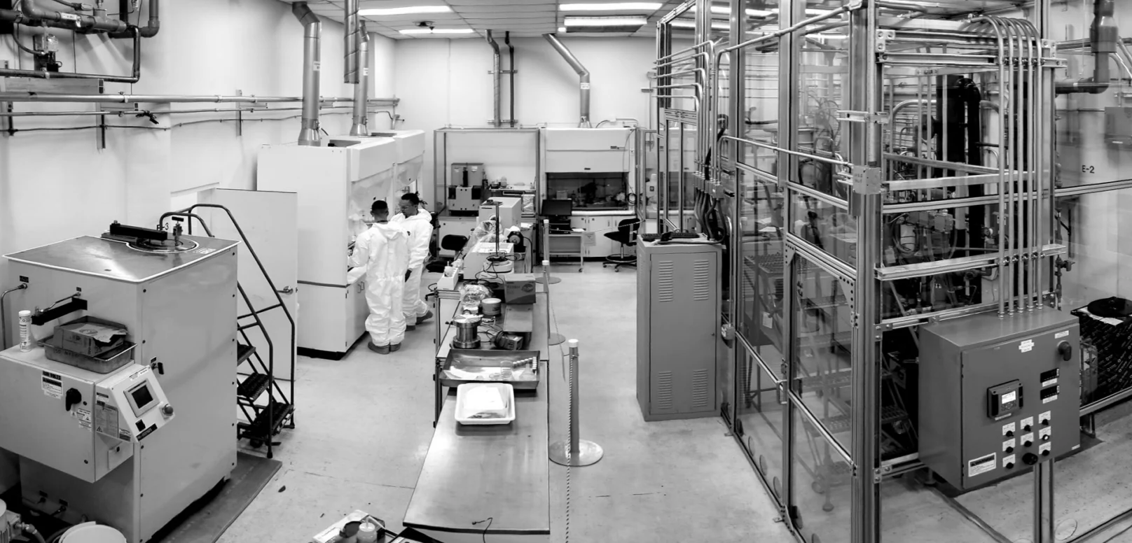 Black and white image of a laboratory with scientific equipment, two people in protective suits working at a station, and various machines and cabinets throughout the room.