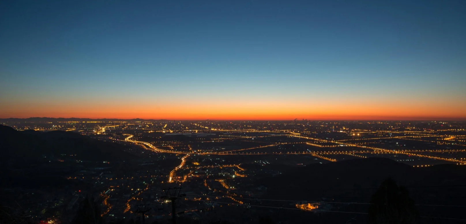 City lights stretch across a flat landscape at dusk, with a clear sky transitioning from deep blue to orange near the horizon.