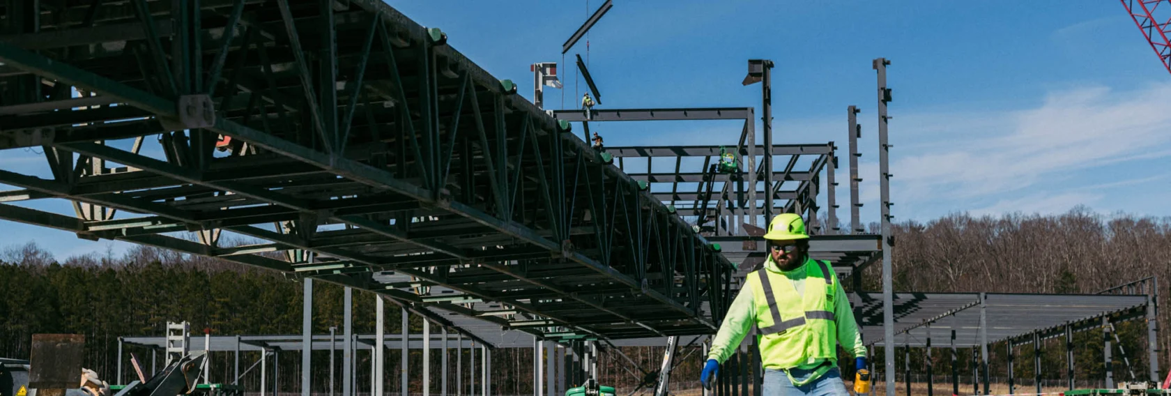 A construction worker in a high-visibility jacket and helmet walks on a building site with steel framework and cranes in the background.