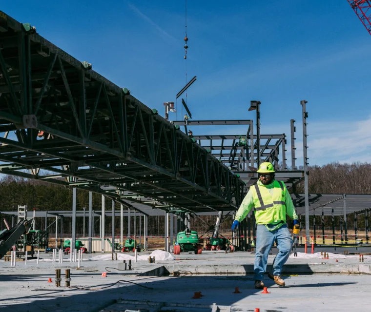 A construction worker in a high-visibility jacket and helmet walks on a building site with steel framework and cranes in the background.
