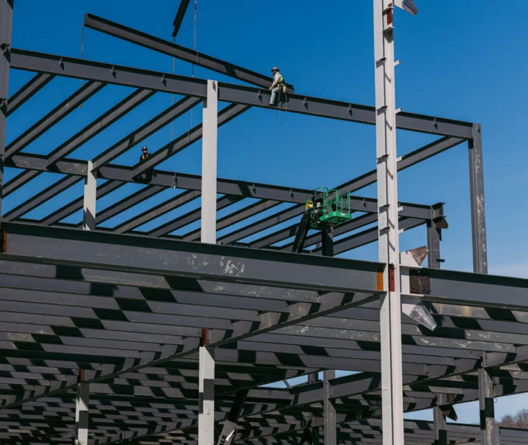 A construction worker sits on a steel beam of a building framework while a crane hoists another beam into place against a clear blue sky.