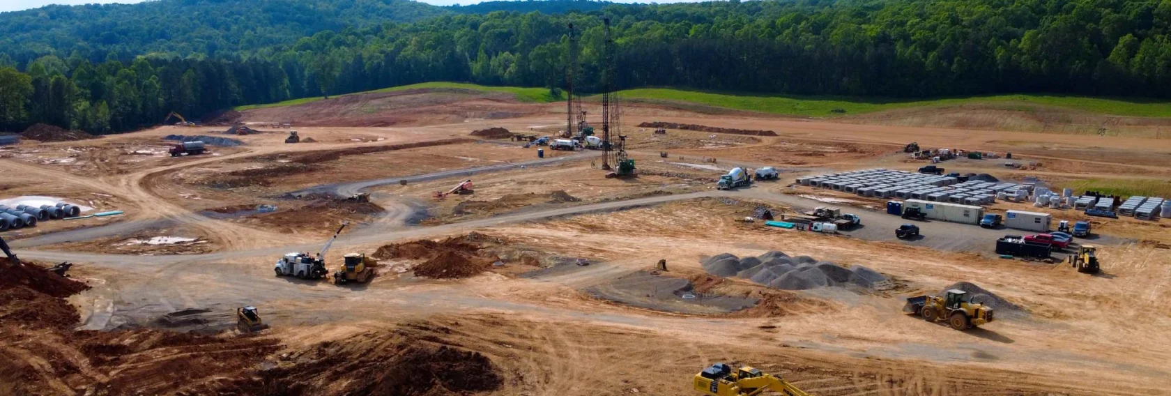 A large construction site with heavy machinery, dirt roads, and materials spread out, surrounded by green hills and trees under a partly cloudy sky.