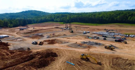 A large construction site with heavy machinery, dirt roads, and materials spread out, surrounded by green hills and trees under a partly cloudy sky.
