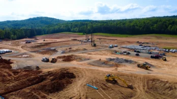 A large construction site with heavy machinery, dirt piles, scattered materials, and a forested hill in the background under a partly cloudy sky.