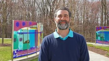 A man with a beard stands outside near colorful glass panels and leafless trees on a sunny day.