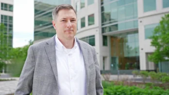 A man in a gray checked blazer and white shirt stands outside a modern glass office building with greenery in the background.