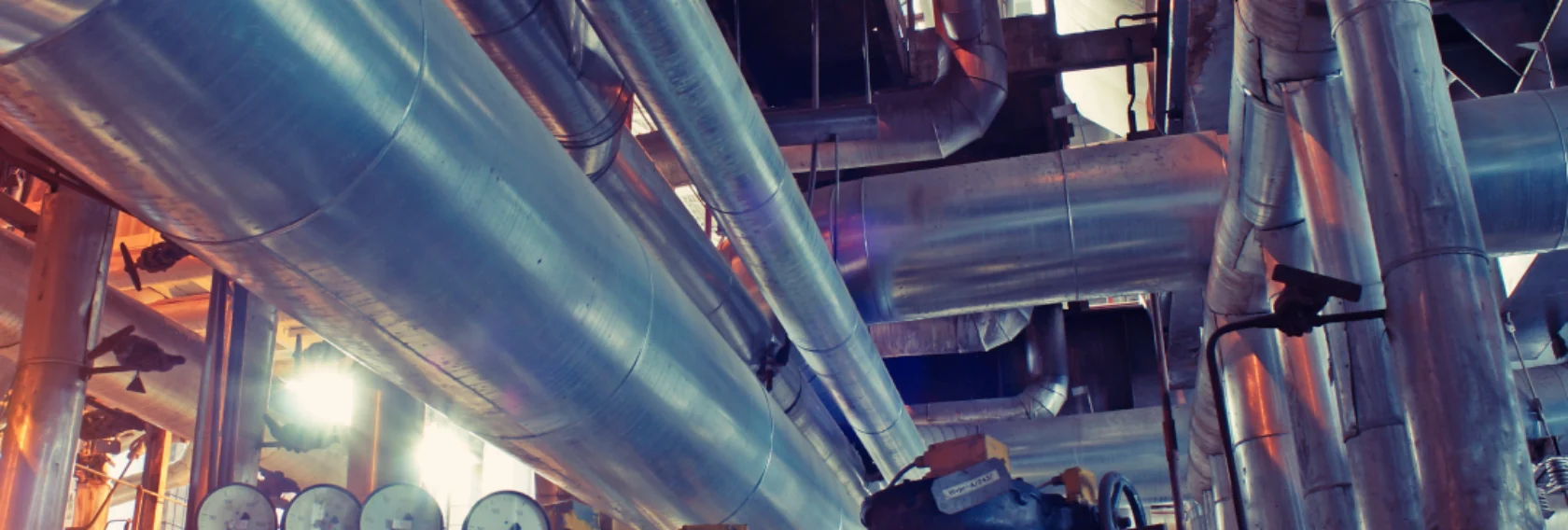 Interior of an industrial facility with large metal pipes, pressure gauges, and control valves visible throughout the space.