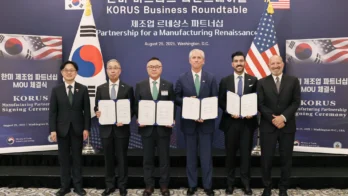 Six men in business attire stand side by side holding signed agreements at the KORUS Manufacturing Partnership Signing Ceremony in Washington, D.C., with flags and a presentation banner behind them.