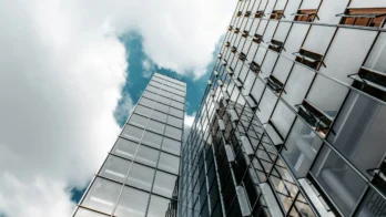 Glass skyscrapers with reflective windows rise toward a partly cloudy sky, capturing cloud and sky reflections on their surfaces.