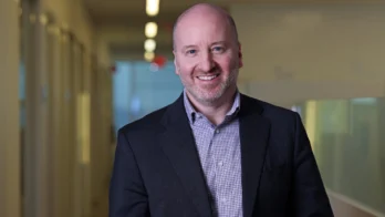 A man in a suit jacket and checked shirt stands smiling in a hallway with blurred office windows and lights in the background.