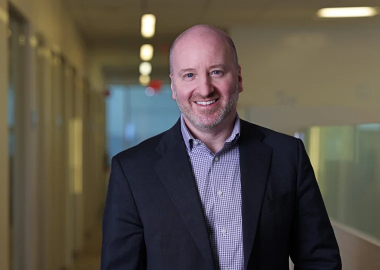 A man in a suit jacket and checked shirt stands smiling in a hallway with blurred office windows and lights in the background.