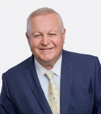 A middle-aged man with short gray hair in a blue suit, light blue shirt, and patterned yellow tie, smiling against a plain light background.