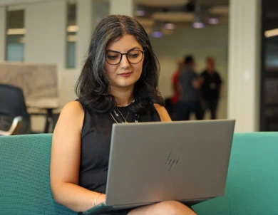 A woman with glasses sits on a green couch, working on an HP laptop in a modern office setting.