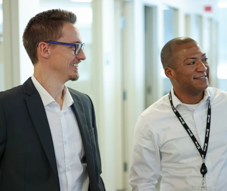 Two men in business attire stand side by side in an office hallway, smiling and looking in the same direction.