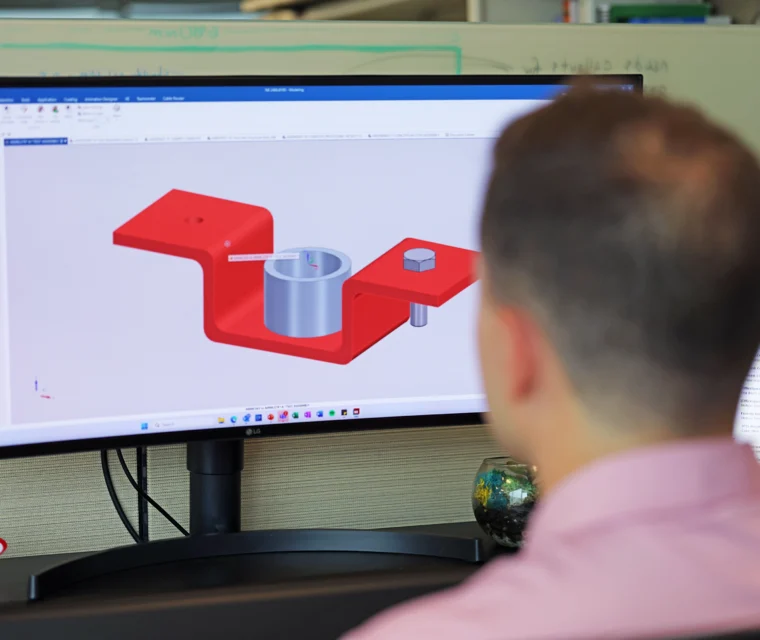 A person views a 3D CAD model of a red mechanical bracket with a cylindrical part and a bolt on a computer monitor in an office setting.