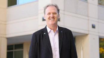 A man in a dark blazer and light shirt stands smiling in front of a modern building with large windows.