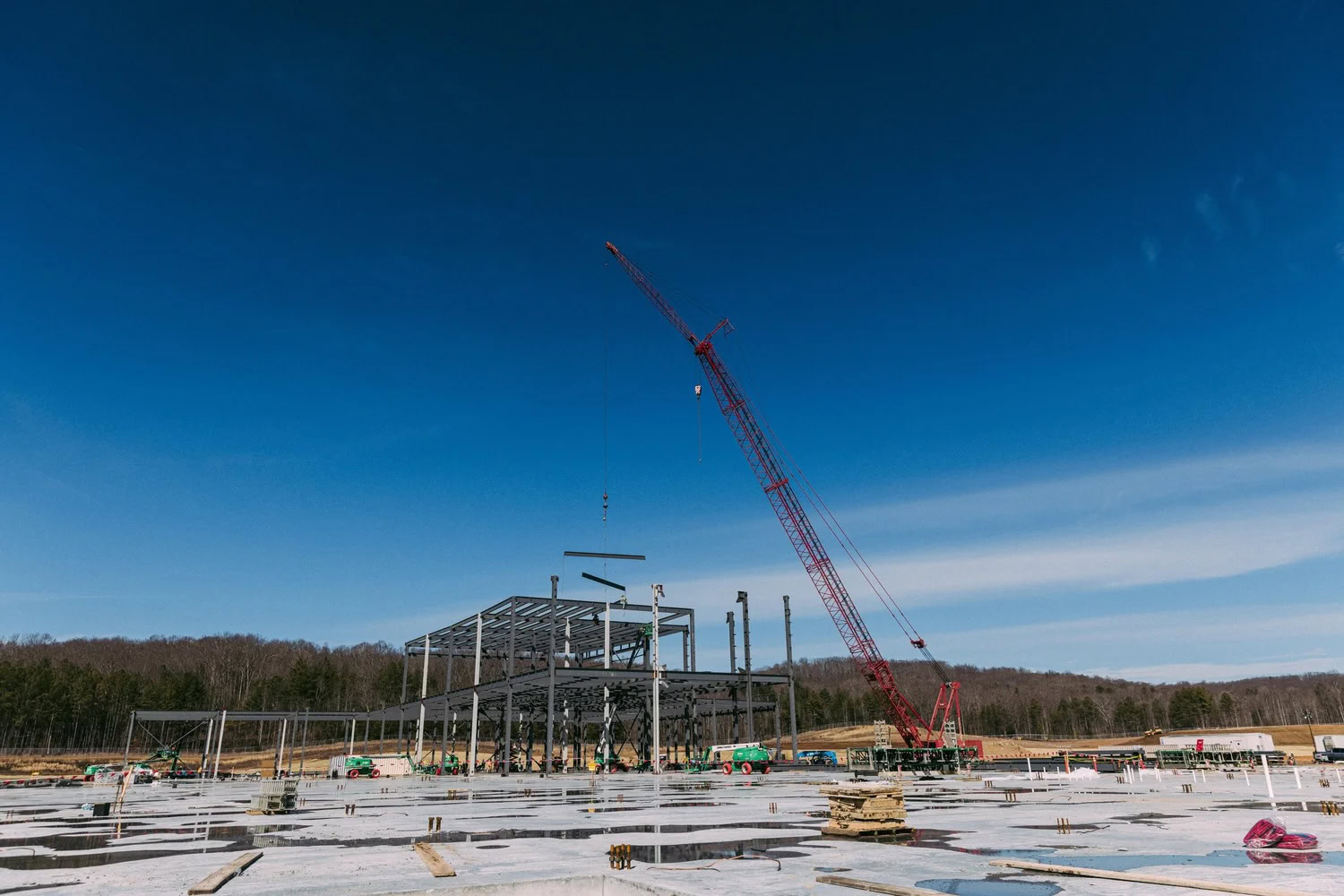 A red crane lifts steel beams at a construction site with a partially built metal framework under a clear blue sky.