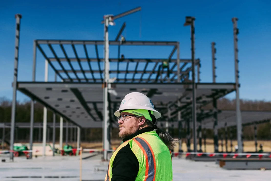 Person in a safety vest and hard hat stands at a construction site with a steel building framework in the background under a clear sky.