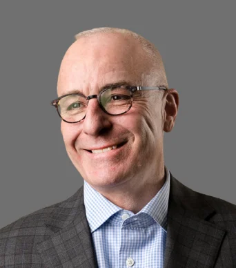 A middle-aged man with glasses, short gray hair, and a checked suit jacket over a blue checkered shirt, smiling against a plain gray background.