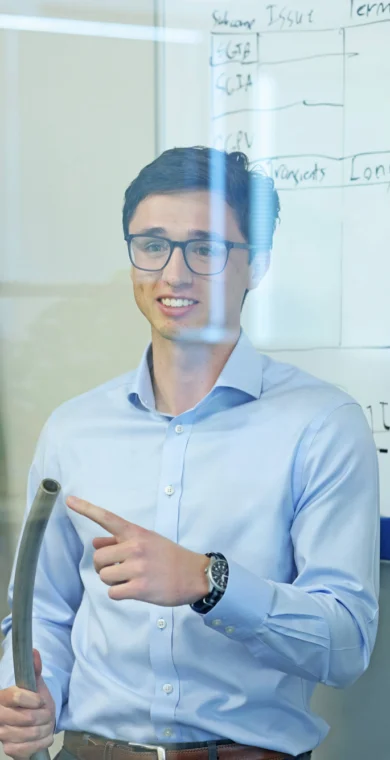 A man in glasses stands in front of a whiteboard with diagrams, holding a metal pipe and pointing, wearing a light blue shirt in an office setting.